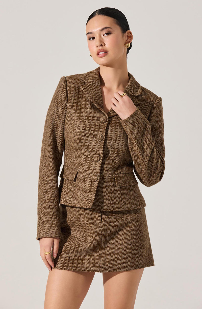 A woman is wearing a matching brown herringbone tweed blazer and skirt. The blazer is tailored with a notched lapel collar, three buttons down the front, and flap pockets on each side. The skirt is short and straight, matching the blazer's pattern. She has dark hair pulled back, gold earrings, and is accessorizing with gold rings.