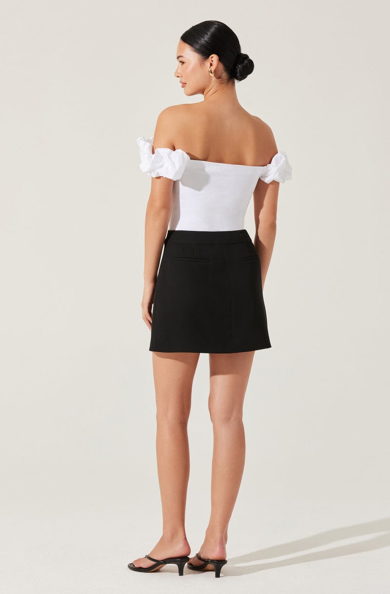 A woman is wearing an off-the-shoulder white top with ruffled sleeves and a fitted bodice. She pairs it with a high-waisted black skirt that has a straight shape and slit pockets in the back. She completes her outfit with black heeled sandals.