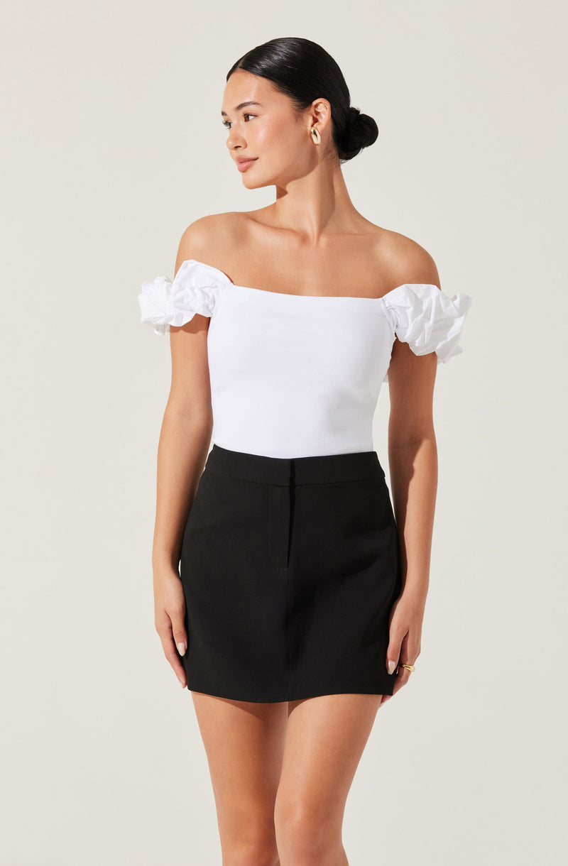 A woman is wearing an off-the-shoulder white top with puffed short sleeves and a fitted bodice, paired with a high-waisted black skirt that falls mid-thigh.
