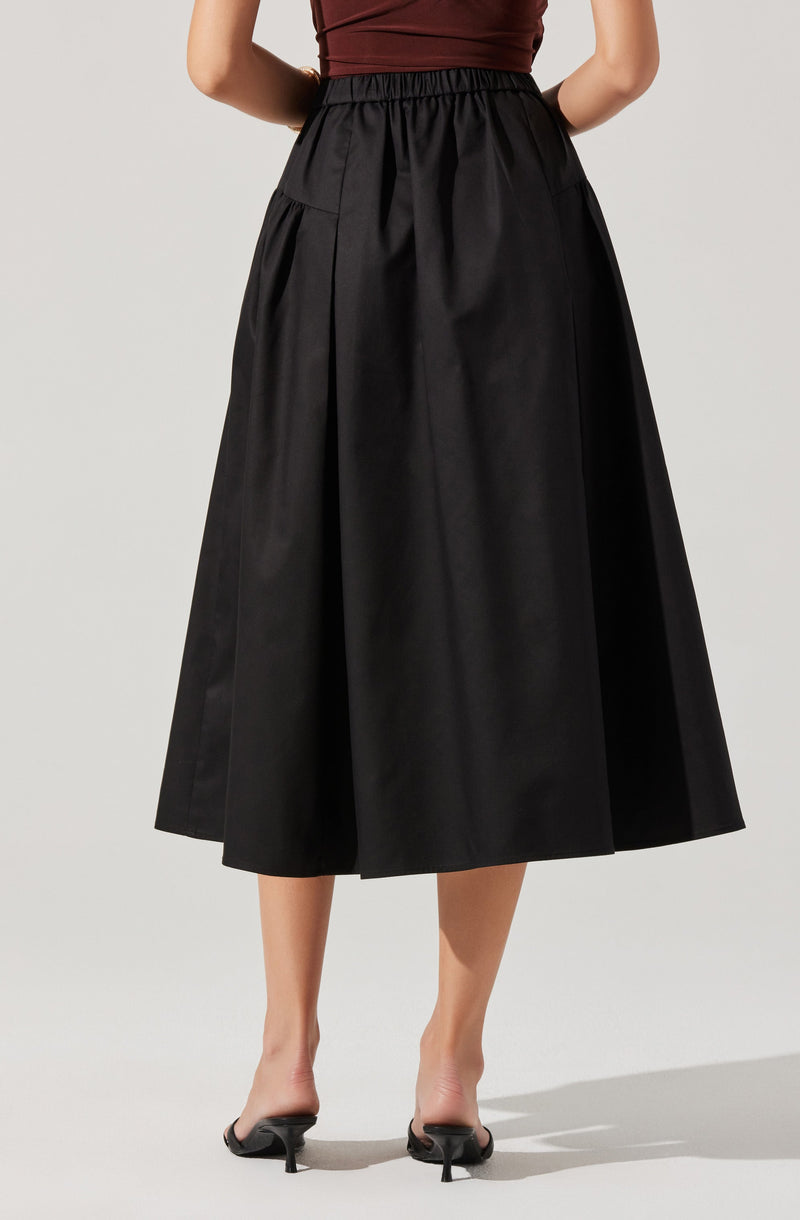 A woman is wearing a high-waisted, ankle-length black skirt with a full, voluminous silhouette. The skirt features an elastic waistband and large front pockets. She is also wearing a fitted, sleeveless top in a dark red or burgundy color.