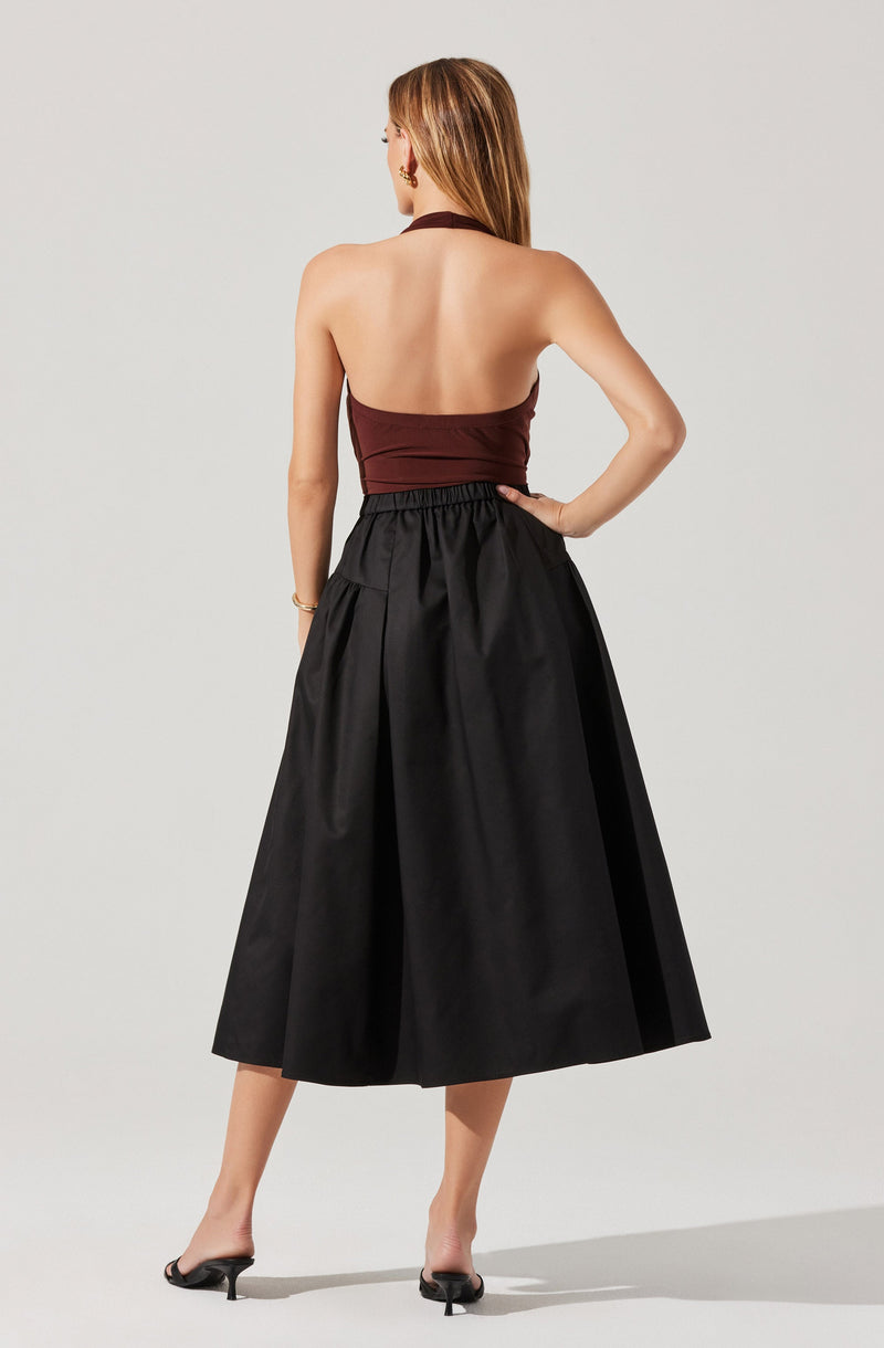 A woman is wearing a sleeveless, halter-style top in a deep burgundy color, which wraps around her neck and ties at the back. She has paired it with a high-waisted black midi skirt that is full and pleated, creating a voluminous silhouette. The skirt falls to mid-calf length. The woman is also wearing black open-toe heels.