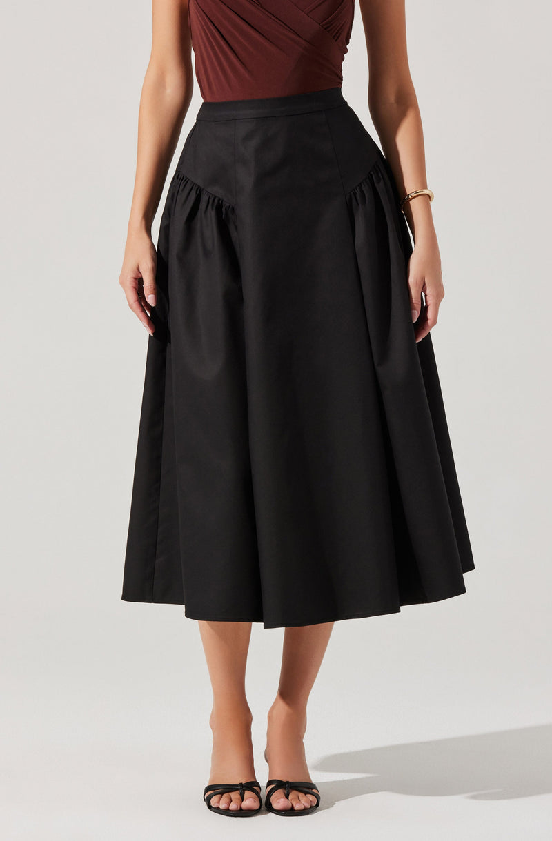 A woman is wearing a sleeveless, fitted, maroon top with a slight drape at the front and a high-waisted, wide-leg black midi skirt with gathered detailing on the sides. She has on black open-toe heels with thin straps and a gold bracelet on her right wrist.
