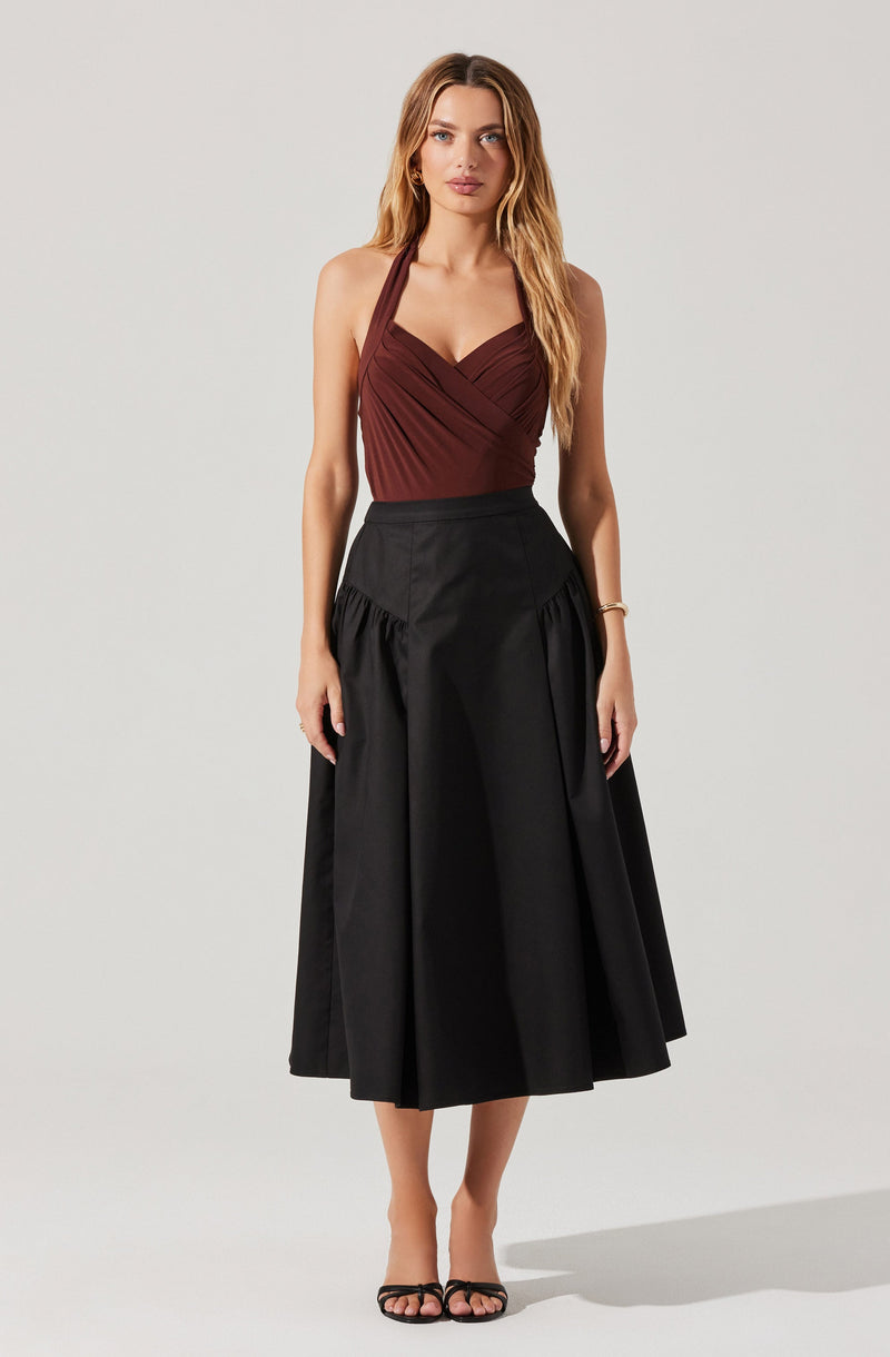 A woman is wearing a sleeveless, burgundy halter top with gathered detailing at the bust, paired with a high-waisted, black midi skirt that features a full, pleated design. She is also wearing black strappy high-heeled sandals.