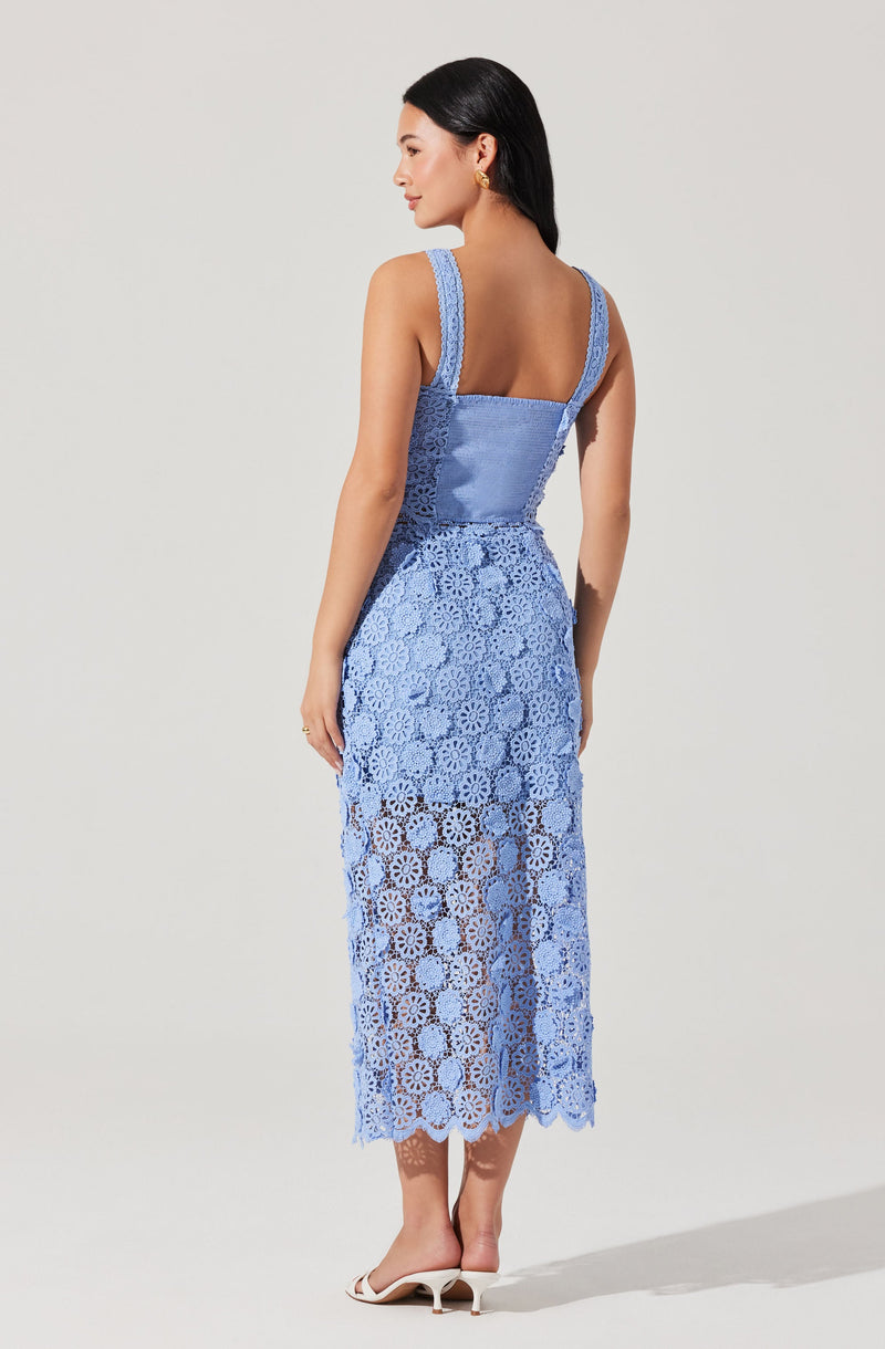 A woman is wearing a sleeveless, light blue crochet dress with a fitted bodice and wide shoulder straps. The dress features intricate crochet patterns throughout, with the skirt portion being semi-sheer and extending to mid-calf length. She is also wearing white heeled sandals.