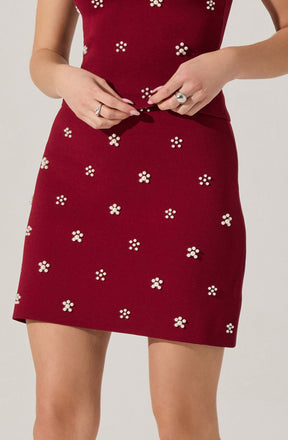 A woman is wearing a red dress decorated with small, white, floral-shaped embellishments. The dress is fitted at the waist and has a straight skirt that falls above the knees. She is adjusting the hem of the dress with her hands.