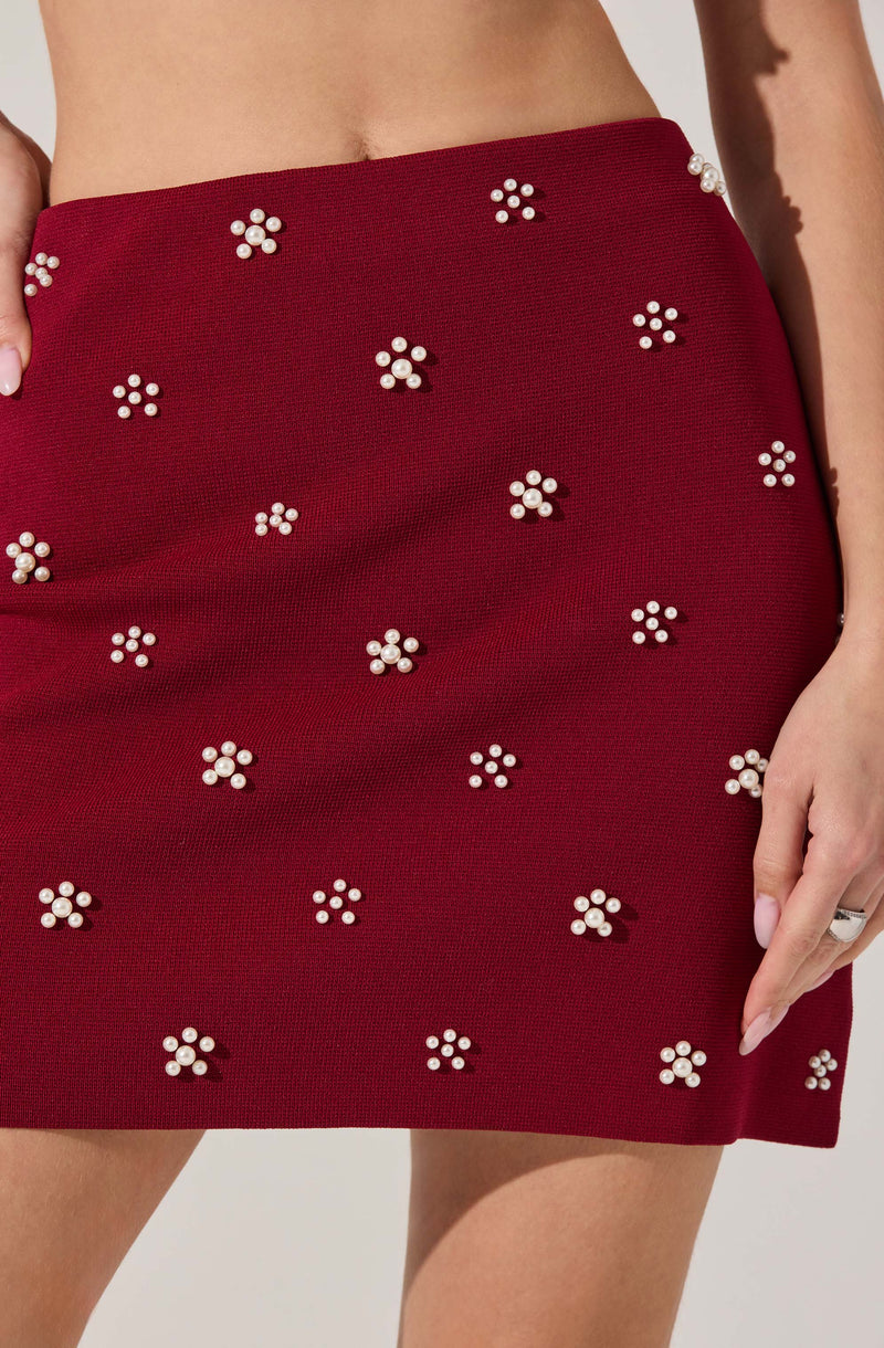 A close-up of a woman's red skirt decorated with small clusters of white pearls arranged in flower shapes. The skirt has a textured fabric and sits high on the waist. The woman's left hand rests on her thigh, with one finger slightly touching the fabric.