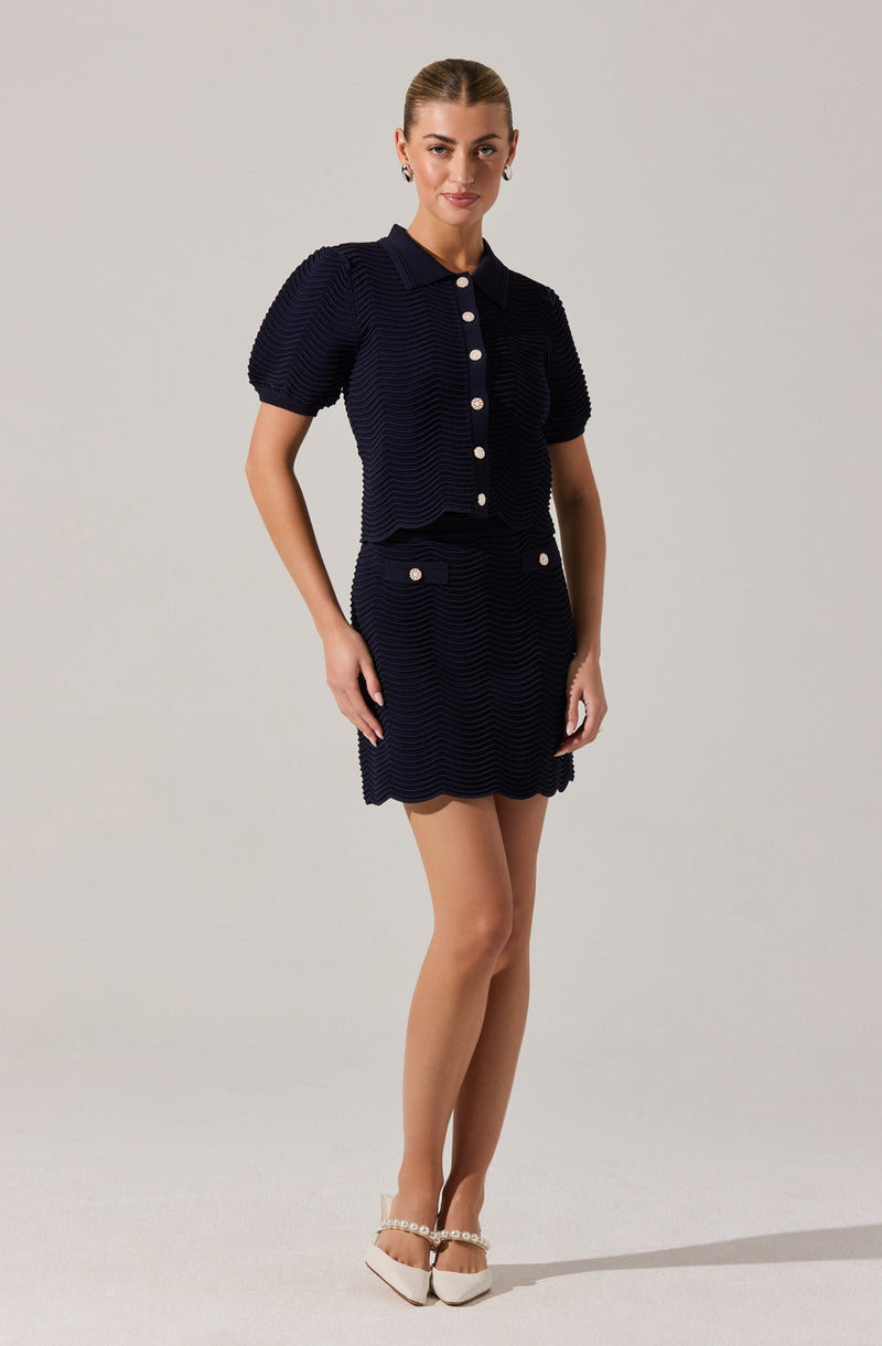 A woman is wearing a matching navy blue textured short-sleeve button-up top with a collar and a matching textured skirt with white buttons and pockets. She pairs the outfit with beige heels decorated with pearl embellishments.
