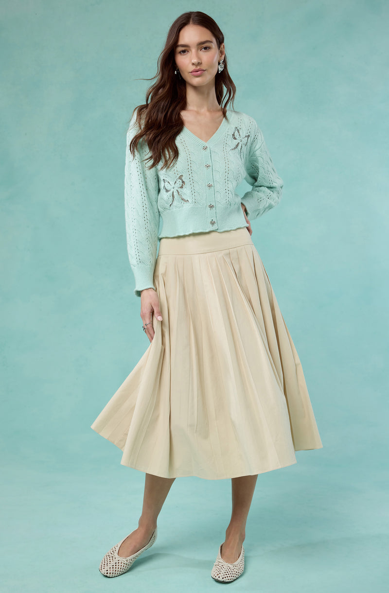 A woman is wearing a light blue, knitted, V-neck cardigan sweater with floral cutout designs on the chest and sleeves, accompanied by small buttons down the front. She is also wearing a long, beige, pleated A-line skirt that reaches mid-calf, paired with white woven flat shoes.