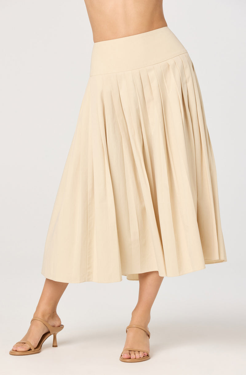 A woman is wearing a high-waisted, cream-colored, pleated midi skirt made of light, flowing fabric. She is also wearing beige open-toe high-heeled sandals with ankle straps.
