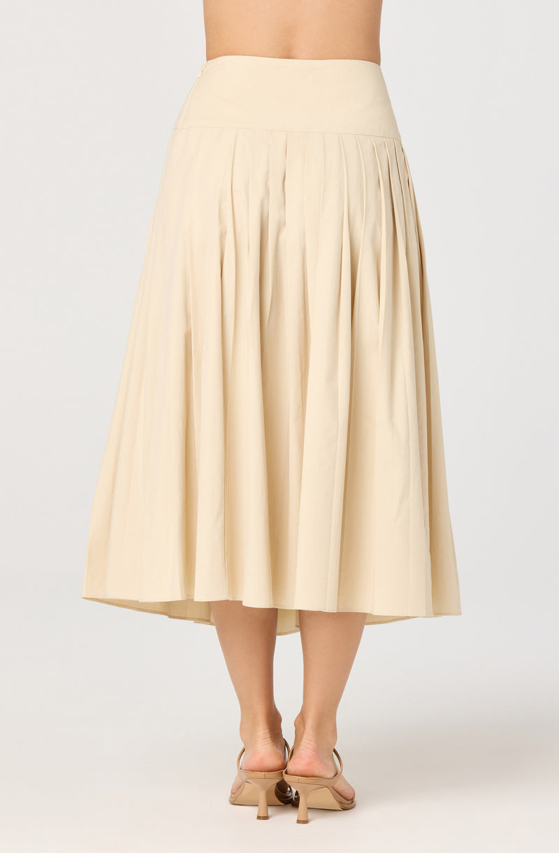 A high-waisted, cream-colored, pleated midi skirt that falls below the knees. The skirt features evenly spaced pleats throughout, creating a flowing and voluminous silhouette. The fabric appears to be lightweight with a smooth texture. The model is wearing nude-colored heels with a small heel and ankle straps.