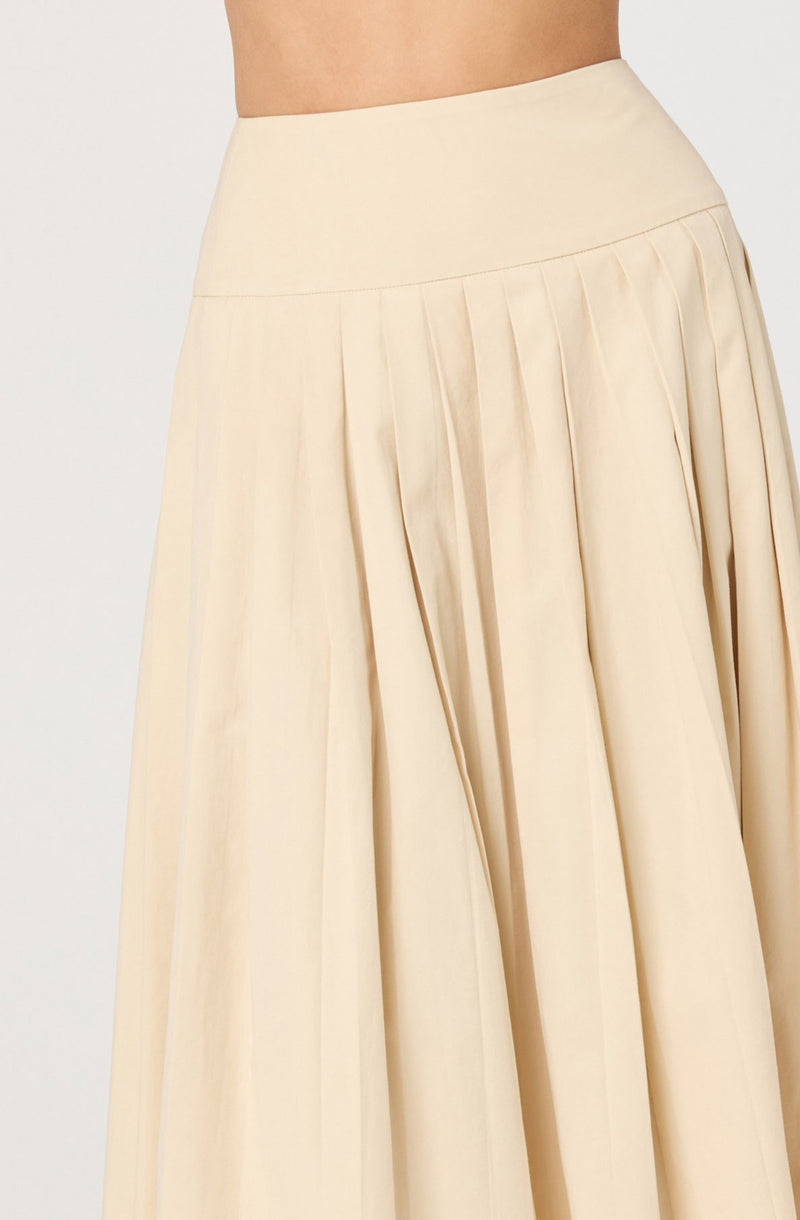 A woman is wearing a high-waisted, floor-length skirt made of a light, creamy-colored fabric. The skirt features a wide waistband and multiple pleats running down from the waist to the hem, creating a flowing and elegant silhouette.