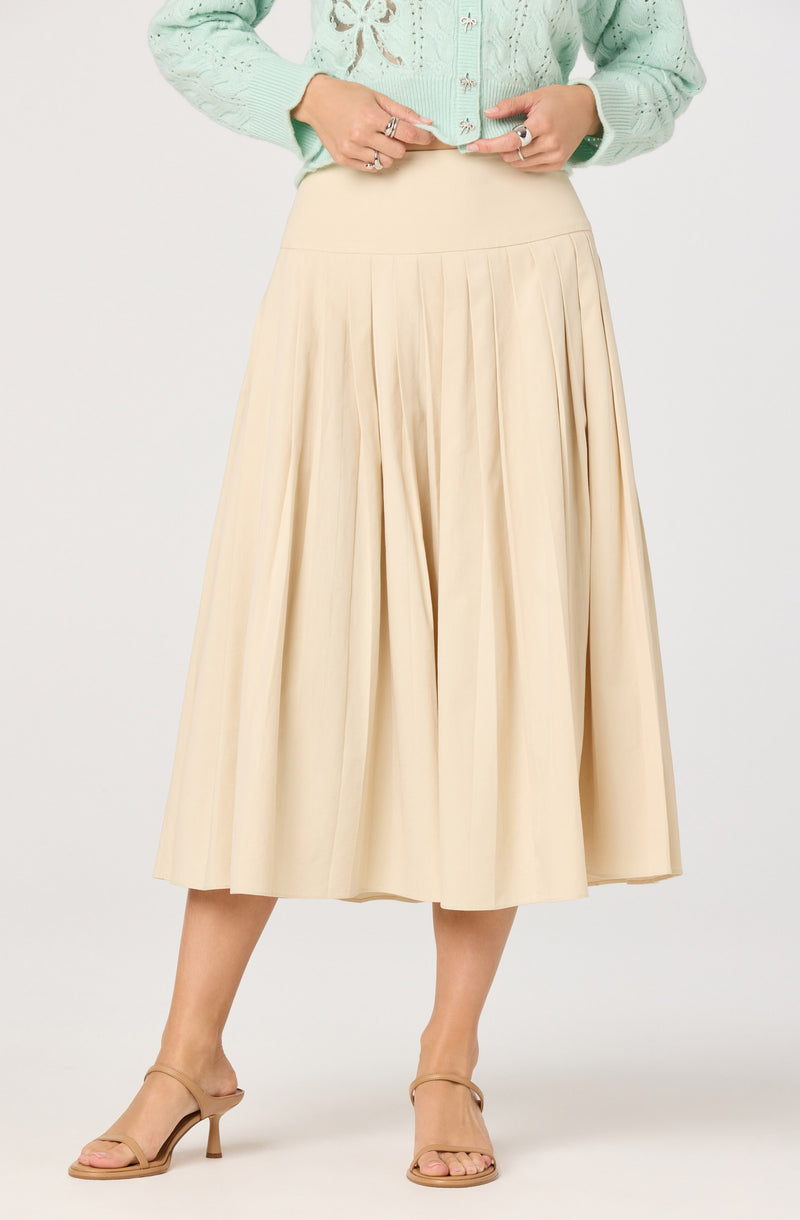 A woman is wearing a light green, long-sleeved, button-up sweater with embroidered details. She is also wearing a high-waisted, cream-colored, pleated midi skirt that falls below her knees. She has on beige strappy high-heeled sandals.