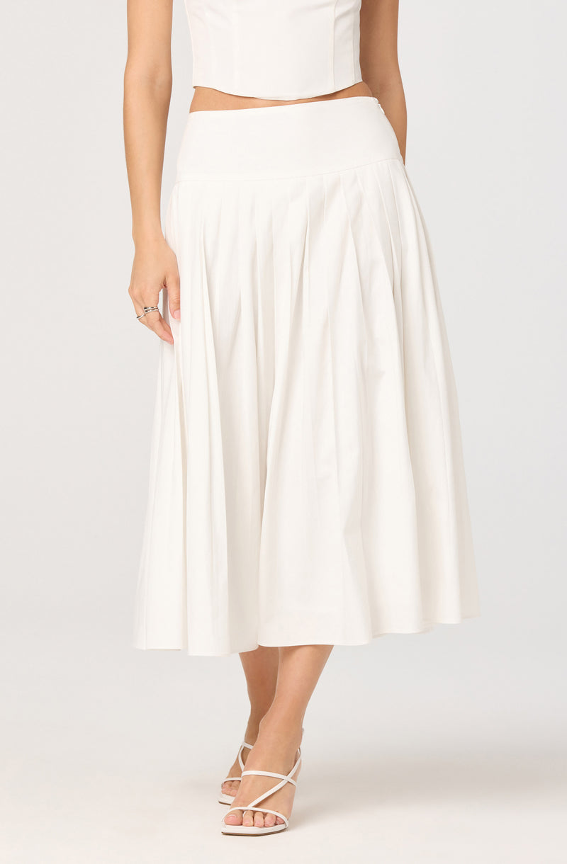 A woman is wearing a high-waisted, ankle-length white skirt with pleats. She has paired it with a sleeveless white crop top, and she is wearing strappy white high-heeled sandals.