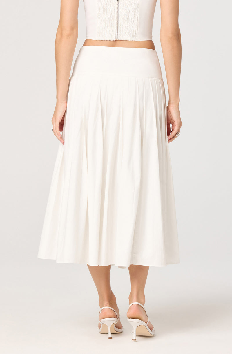 The image shows a woman wearing a high-waisted, ankle-length, white pleated skirt. She is also wearing white high-heeled sandals with thin straps. The top she is wearing is a white sleeveless cropped top with a zipper running down the center.