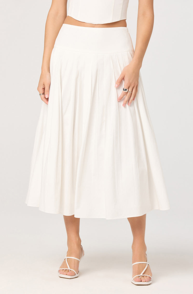 The image shows a woman wearing a high-waisted, cream-colored, pleated midi skirt with a smooth waistband. She is also wearing a sleeveless, cropped white top. The woman has on open-toed, strappy heels in a light color. She has rings on her fingers and her nails are painted a neutral shade. Her arms are relaxed by her sides.