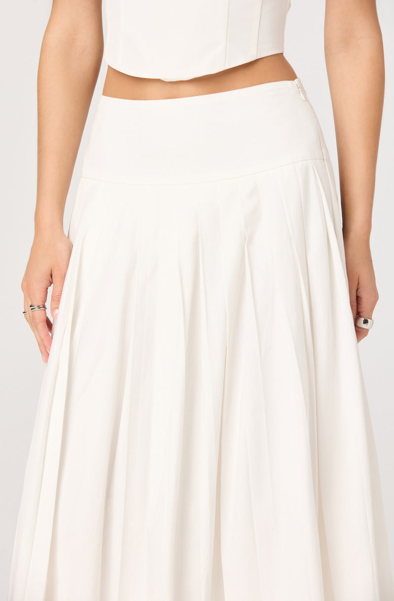 The image shows a person wearing a high-waisted, wide-leg white skirt with pleats, paired with a sleeveless, cropped white top. The skirt appears to be made of a flowing, lightweight fabric with a smooth waistband, and the top is fitted, ending just above the waist. The person is accessorized with rings on their fingers.