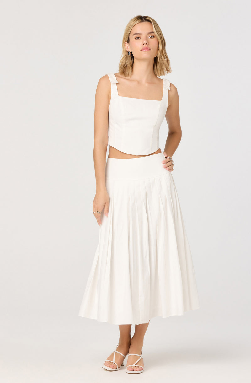 A woman is wearing a white sleeveless crop top with wide shoulder straps that have tied bows and a squared neckline. She is also wearing a white, high-waisted, A-line midi skirt with pleats. She has on white strappy sandals and small hoop earrings.