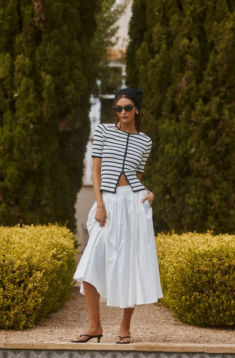 Woman wearing a black and white striped top, black hat, sunglasses, white skirt, and high heels.