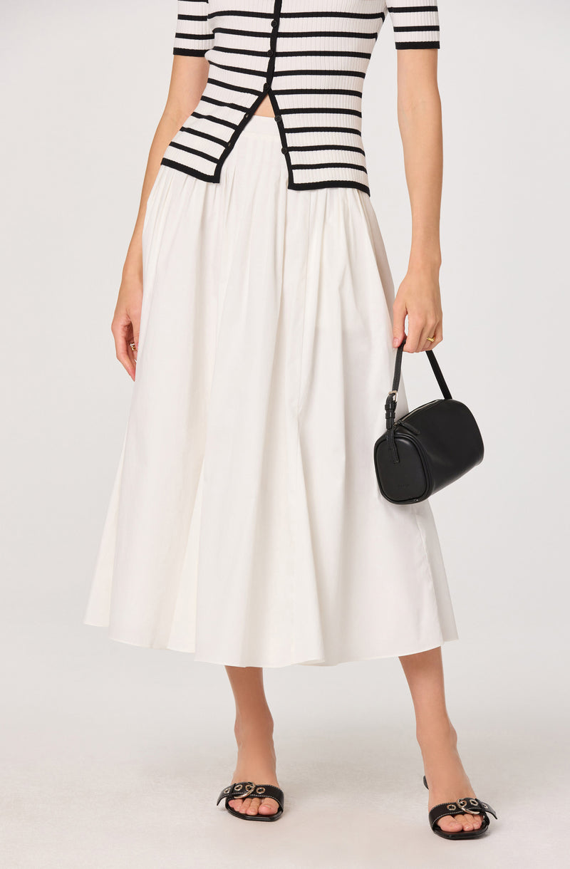 Woman wearing a black-and-white striped short-sleeve top and a white, pleated, midi skirt, holding a black handbag.