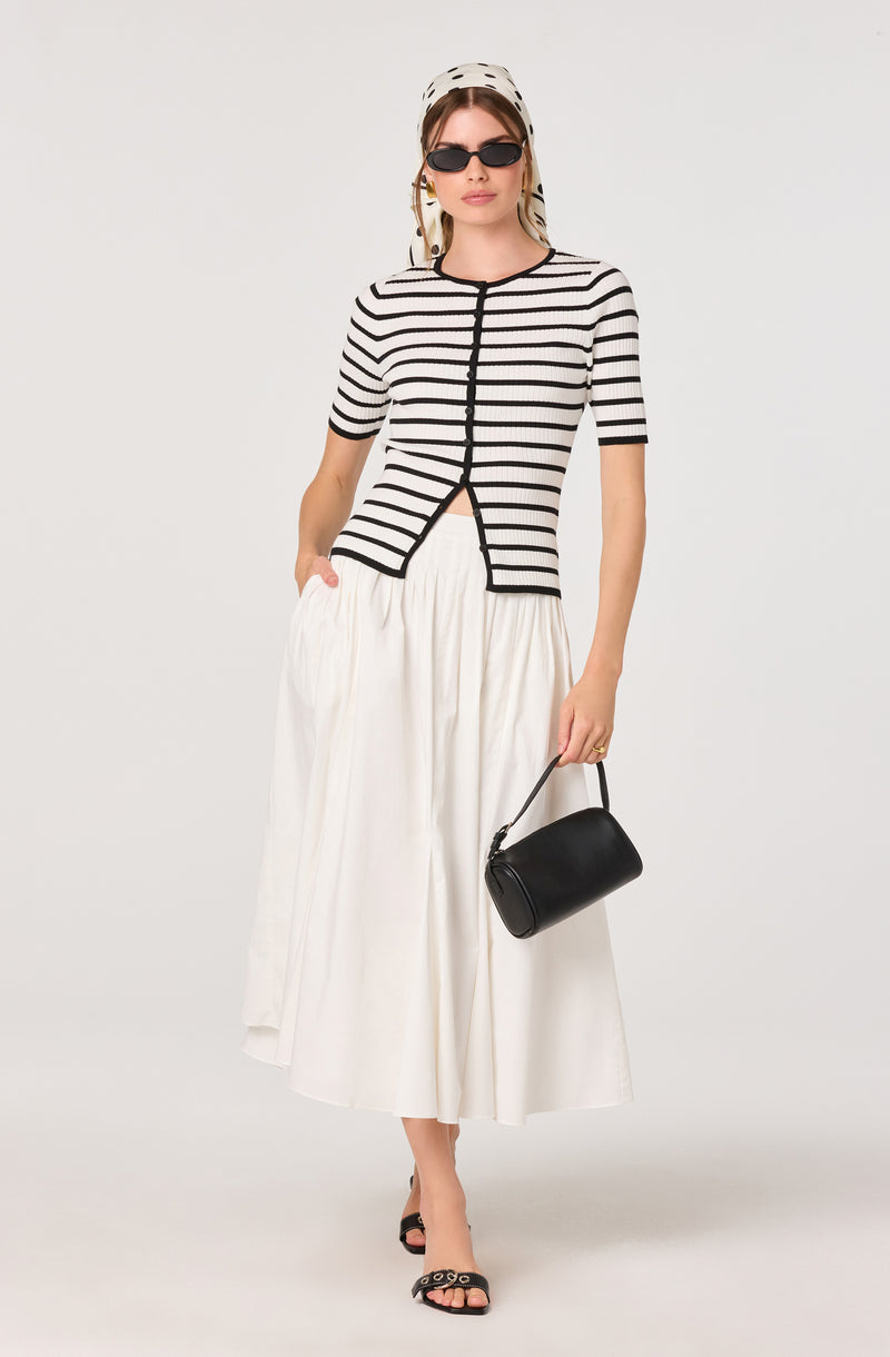 Woman in a black and white striped short-sleeve top, white skirt, black sunglasses, headscarf, black handbag, and black open-toe heels.