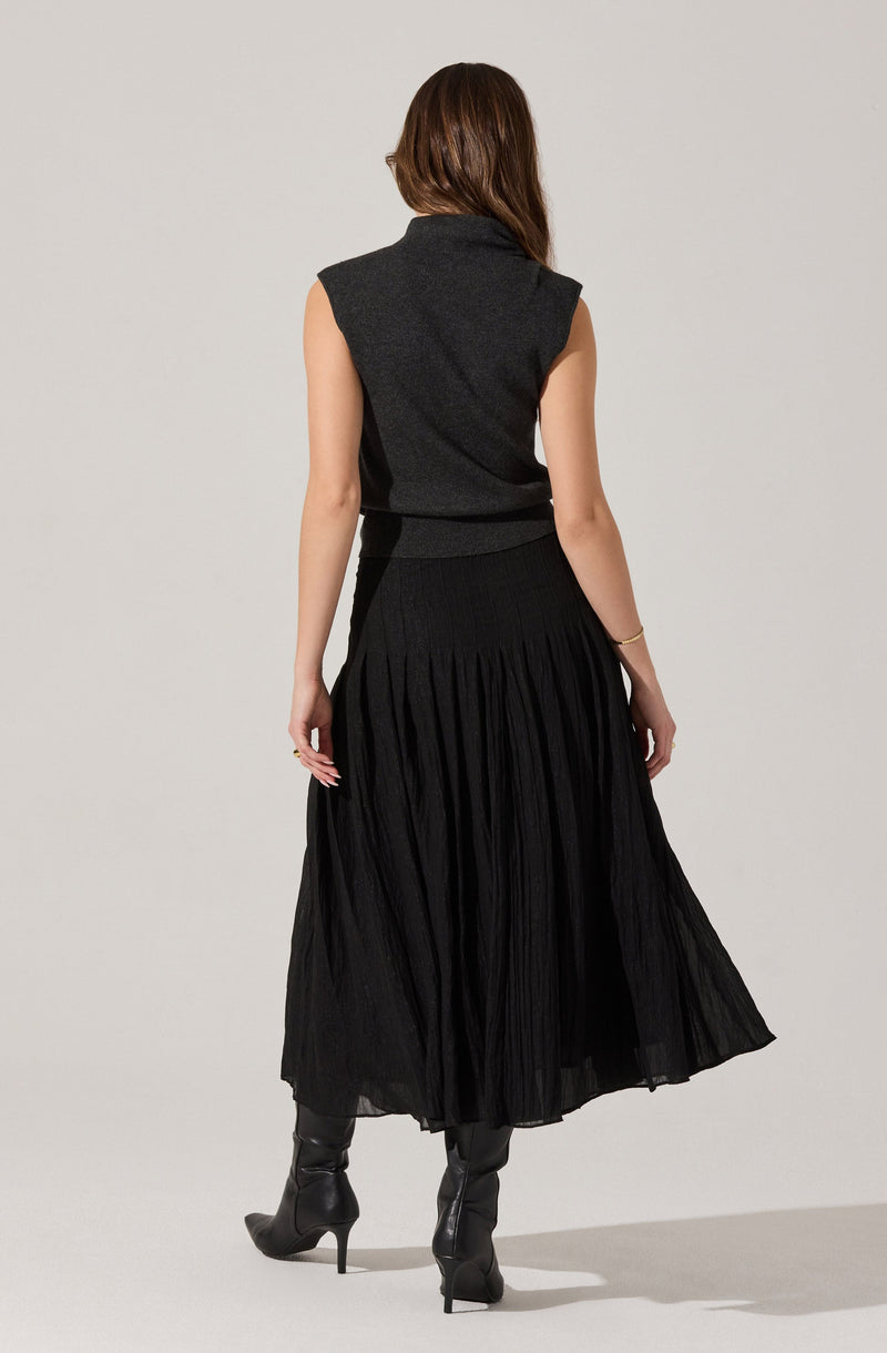 The woman is wearing a dark sleeveless top with a high collar, fitted at the waist. She has on a long, pleated black skirt that reaches her ankles, and she is wearing black high-heeled boots.