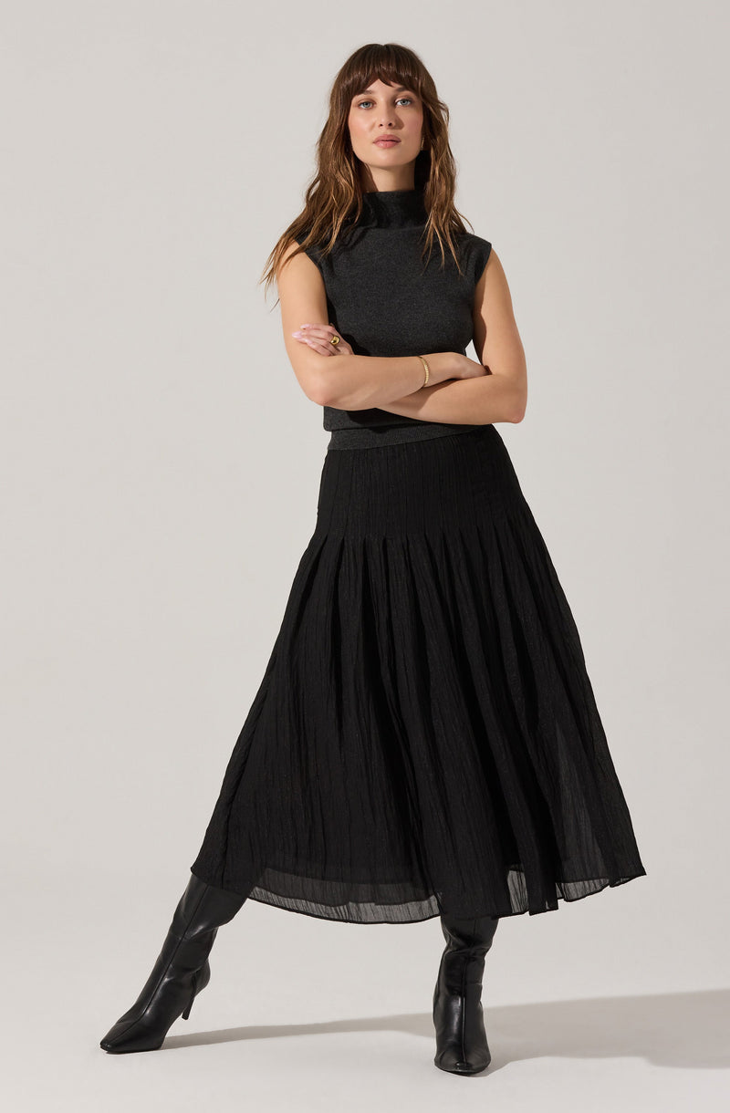 A woman is wearing a sleeveless, dark gray high-neck top paired with a long, black pleated skirt. She has on black knee-high boots and is accessorizing with a gold bracelet and a ring.