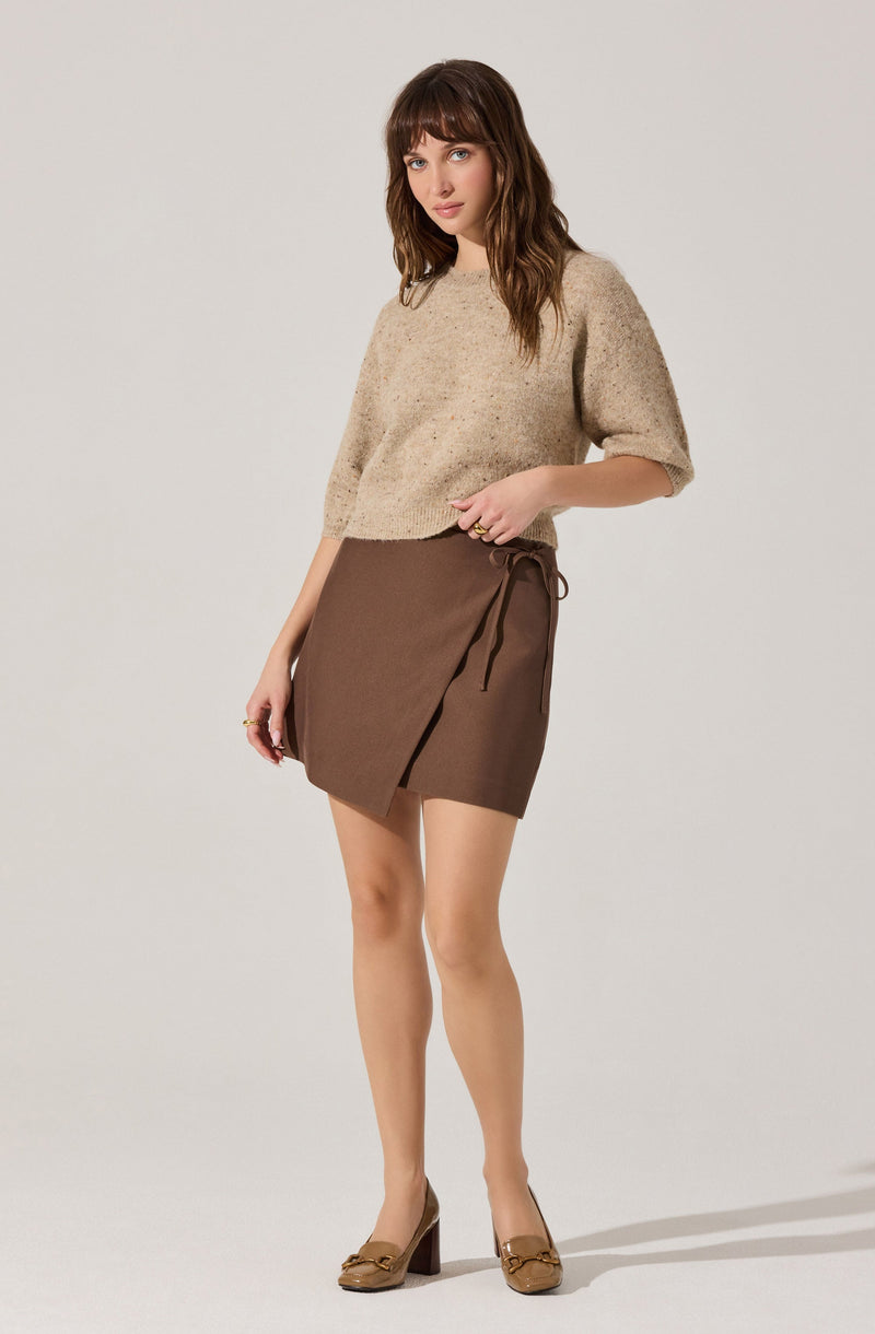 A woman is wearing a beige, textured, loose-fitting sweater with three-quarter length sleeves, paired with a brown wrap-around skirt that features a tie on the side. She is also wearing light brown high-heeled shoes with a decorative element on the front.