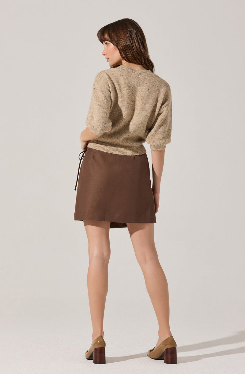A woman is wearing a beige, speckled, short-sleeved knitted sweater with puffed sleeves, paired with a high-waisted, brown A-line mini skirt. She is also wearing beige heeled shoes with chunky wooden heels.
