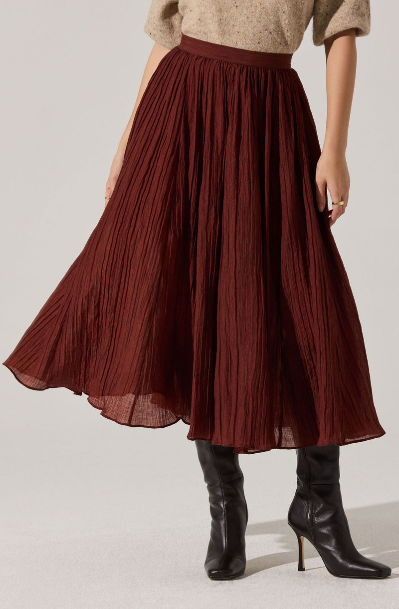A woman is wearing a high-waisted, full-length pleated skirt in a rich, dark red color. The skirt has a textured, slightly sheer fabric with vertical pleats. She pairs it with black knee-high heeled boots and a beige, loose-fitting top with three-quarter sleeves.