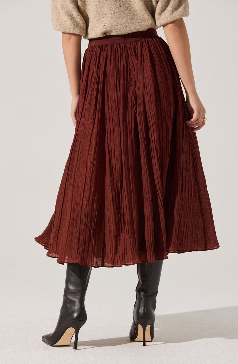 A woman is wearing a high-waisted, ankle-length, pleated skirt made of a textured, rust-colored fabric. She pairs the skirt with a loose, beige sweater and black high-heeled boots.