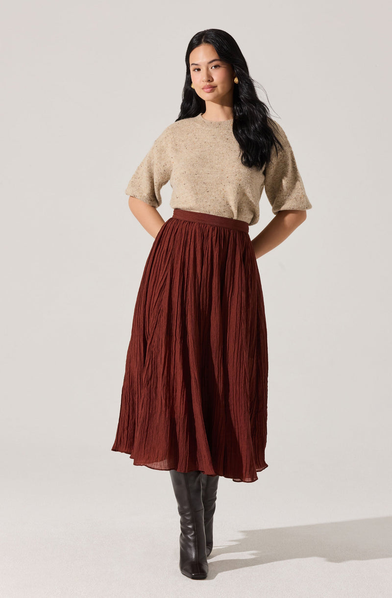 A woman is wearing a beige, short-sleeved, knit sweater with a slightly loose fit. She pairs it with a high-waisted, rust-colored, pleated midi skirt. She completes her outfit with tall, black leather boots.
