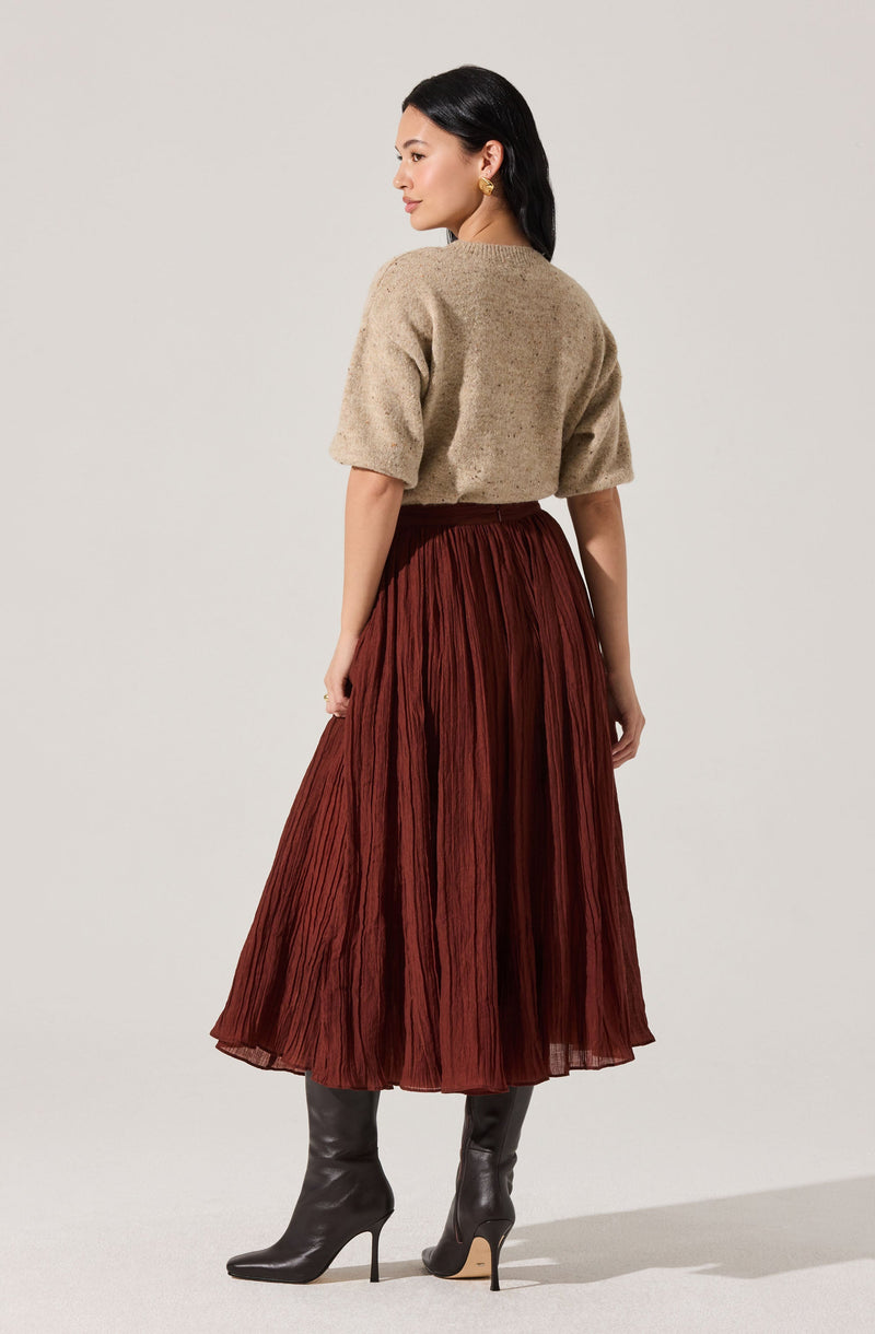 A woman is wearing a beige, short-sleeved, textured knit sweater paired with a high-waisted, pleated, rust-colored midi skirt. She completes the outfit with tall, dark gray leather high-heeled boots.