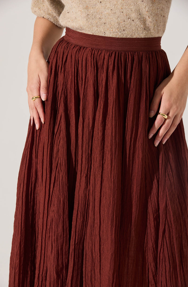 A person is wearing a high-waisted, ankle-length skirt made of crinkled, textured fabric in a rich rust color. The skirt has a wide waistband and vertical pleats that create a flowy silhouette. The person is also wearing a speckled beige top with slightly puffed sleeves. They have both hands resting at their sides, each with a gold ring on their fingers.