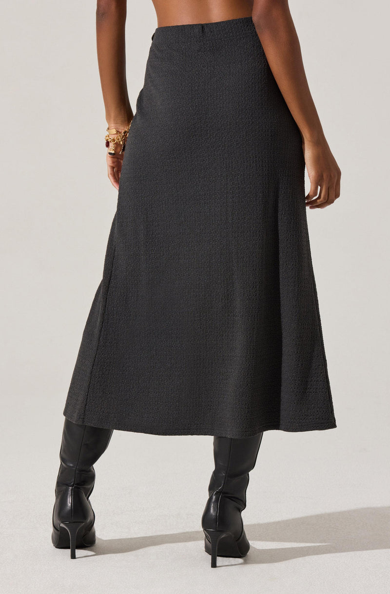 A woman is wearing a long, high-waisted, textured black skirt that reaches her mid-calf. She has on black, high-heeled leather boots. On her left wrist, she is wearing multiple gold bracelets.