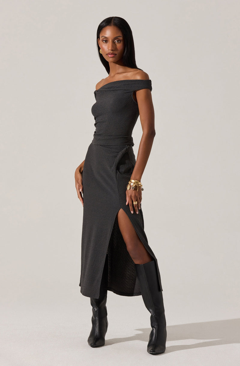 A woman is wearing a fitted, off-the-shoulder black dress with a textured fabric. The dress has a high slit on one side, revealing her leg and knee-high black leather boots. She accessorizes with multiple gold bracelets on her wrist.