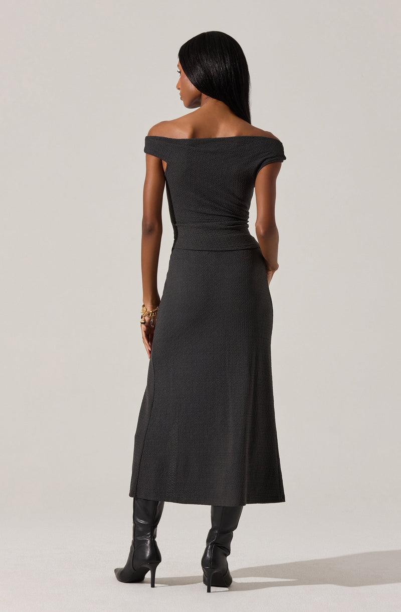 A woman is wearing an off-the-shoulder, fitted black dress with a textured fabric. The dress extends down to her ankles and has a slit on one side. She is also wearing black high-heeled boots and a gold bracelet on her right wrist.