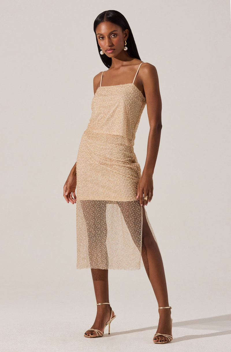 A woman is wearing a beige, sparkly, sleeveless dress with thin spaghetti straps. The dress has a fitted silhouette with a sheer, sparkly overlay starting below the hips and extending to mid-calf, creating a layered look. She is also wearing beige high-heeled sandals with ankle straps.