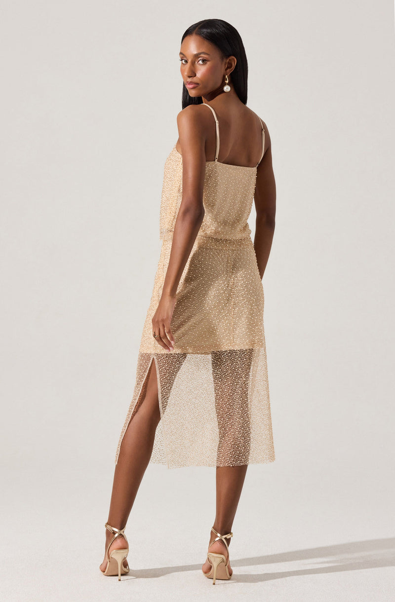 A woman is wearing a sleeveless, beige dress with thin spaghetti straps. The dress has a layered design, featuring a solid upper layer and a see-through, dotted lower layer that extends below the knee. There is a slit on the side of the lower layer, revealing her leg. She is also wearing high-heeled shoes and statement earrings.