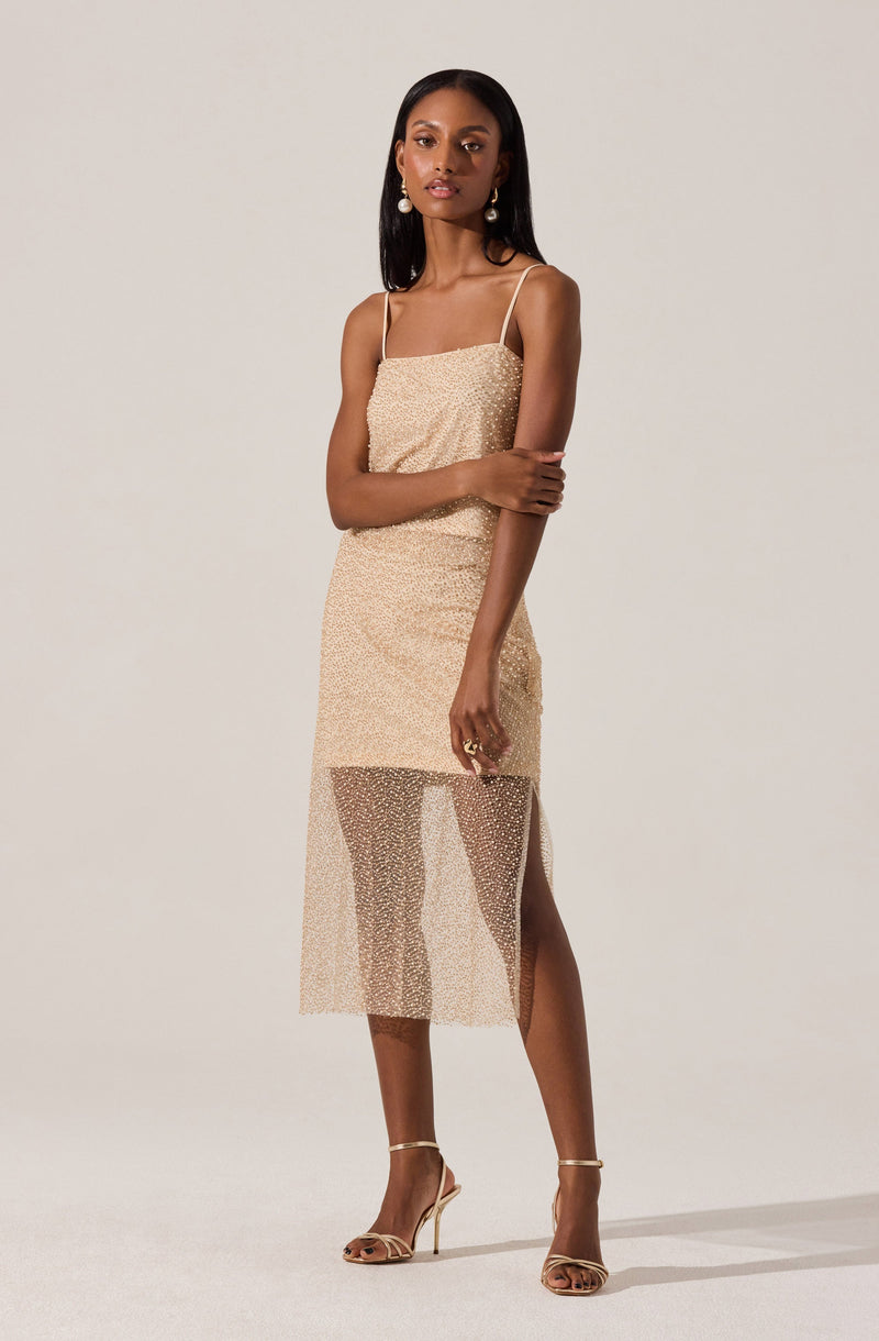 A woman is wearing a beige sleeveless dress with thin spaghetti straps. The top of the dress is fitted and made of a textured fabric, while the skirt features a sheer overlay that extends below the knee with a slit on one side. She is accessorized with large pearl earrings and beige high-heeled sandals with thin straps.
