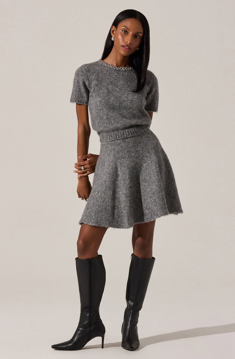 The woman is wearing a matching gray knit sweater and skirt set. The sweater has short sleeves and a round neckline decorated with a row of embellishments. The skirt is flared and falls above the knee. She is also wearing tall black leather boots with high heels.