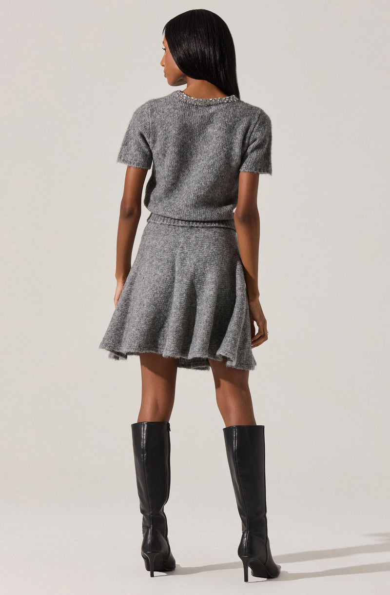 A woman is wearing a short-sleeved, gray knit sweater with a round neckline adorned with small embellishments. She pairs it with a matching gray knit skirt that has a flared, A-line silhouette and a subtle textured pattern. She completes the outfit with tall, black leather knee-high boots that have a pointed toe and stiletto heels.