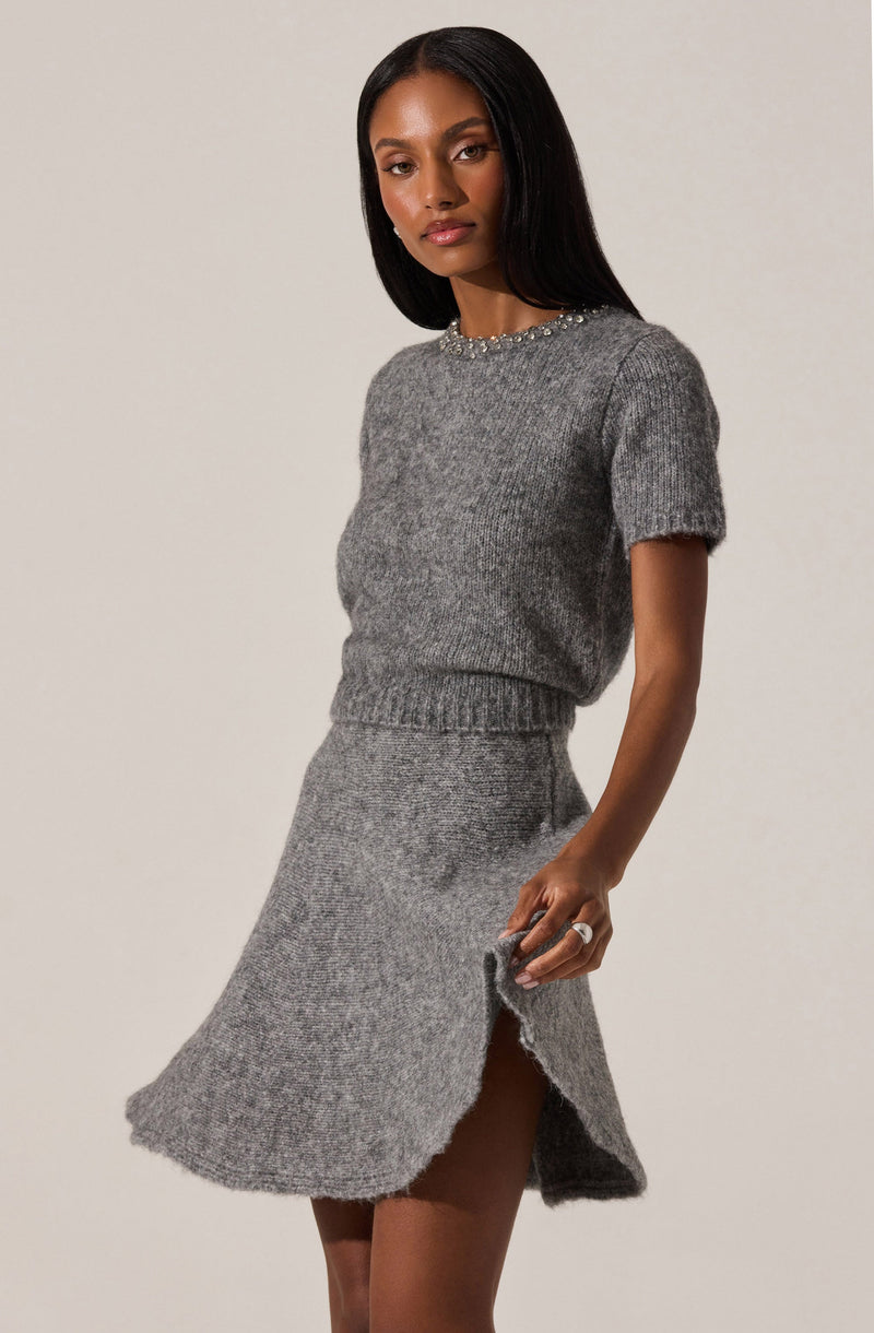 A woman is wearing a matching gray knit sweater and skirt set. The sweater has short sleeves and is embellished with pearls around the neckline. The knit skirt is high-waisted with a side slit, revealing her leg. She has dark, straight hair and a neutral expression.