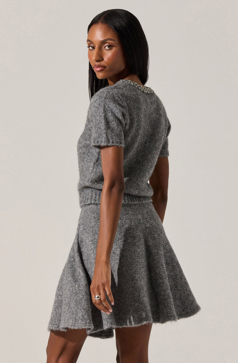 A woman is wearing a gray knit outfit consisting of a short-sleeved, textured sweater with a rounded neckline embellished with small, shiny beads, and a matching high-waisted, flared skirt made of the same knit fabric.