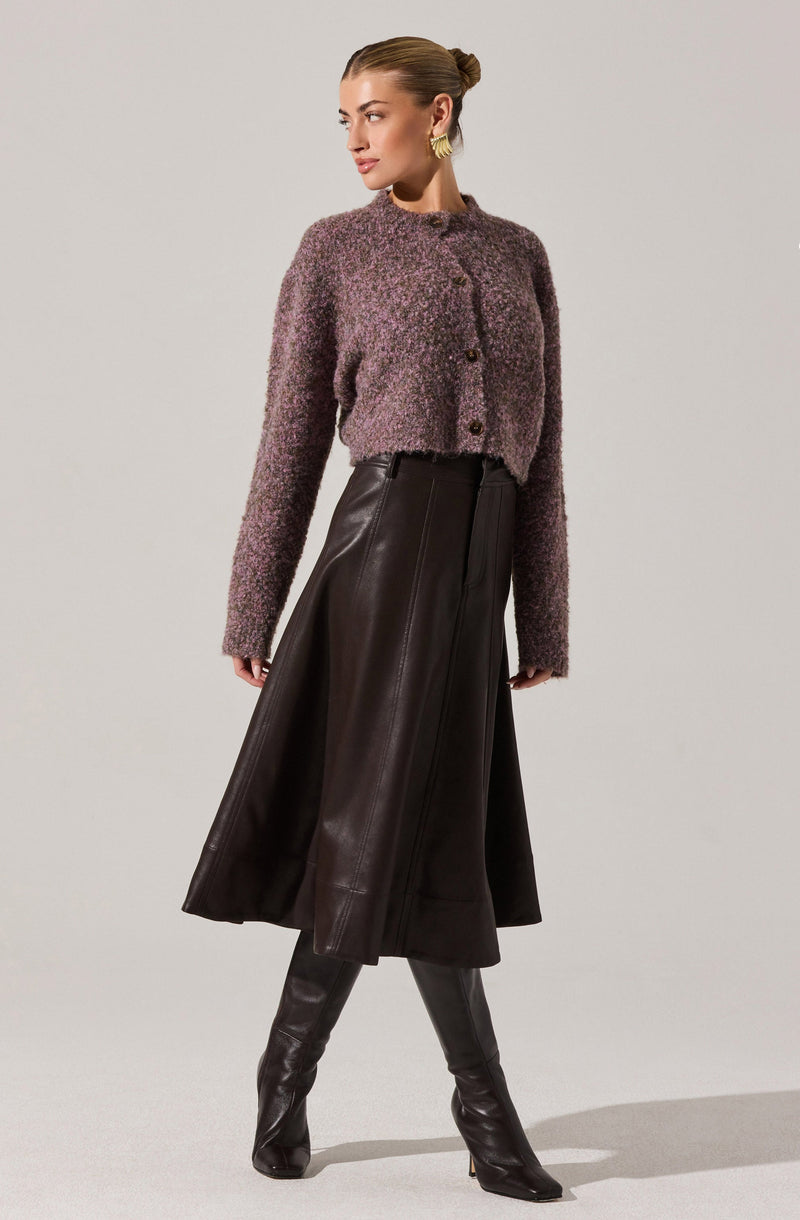 A woman is wearing a textured, cropped, long-sleeve cardigan in a purple and gray mix, with a rounded neckline and buttons down the front. She pairs it with a high-waisted, dark brown, leather midi skirt that flares out slightly, and matching thigh-high leather boots. She is accessorized with large, gold hoop earrings and has her hair styled in a sleek, low bun.