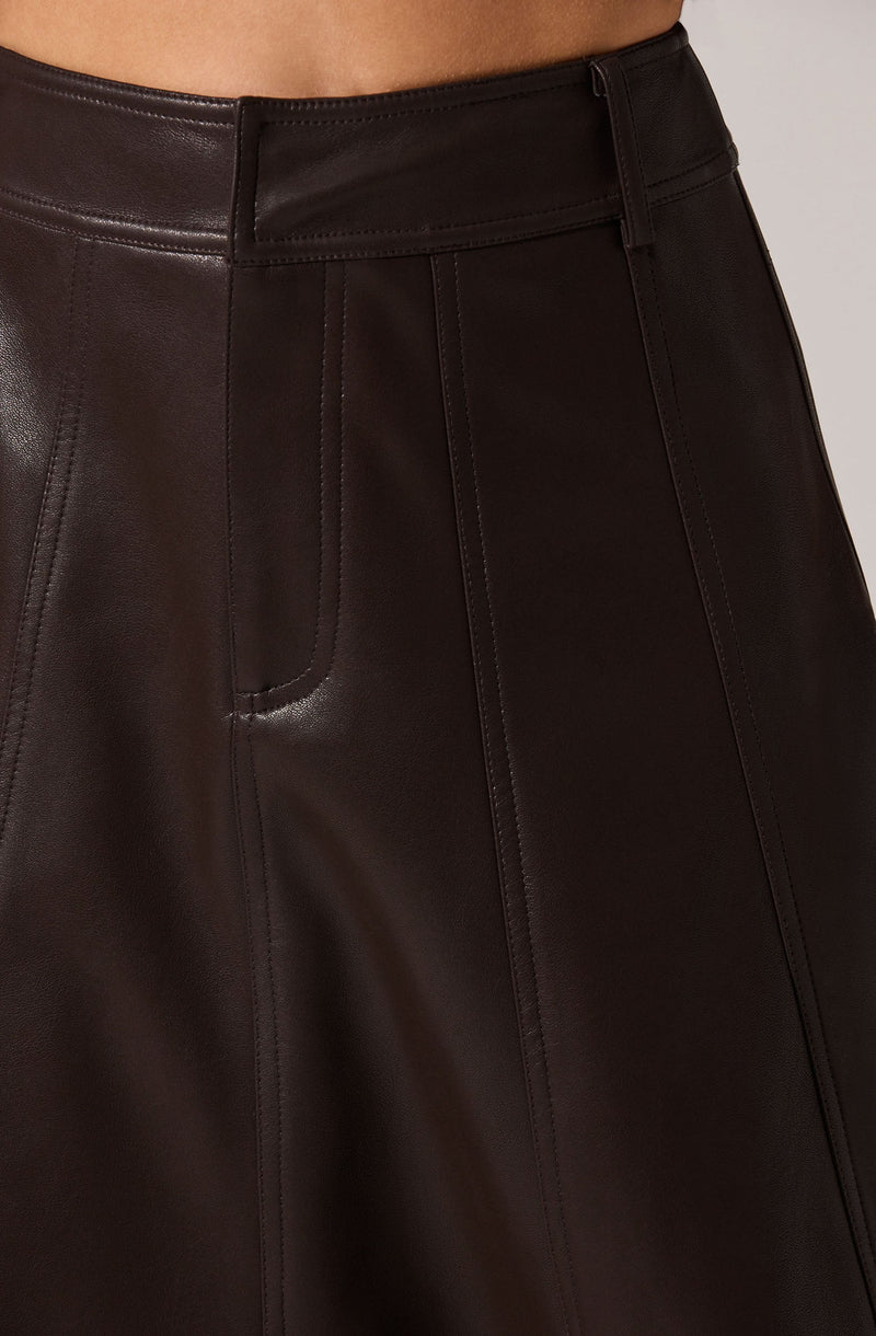 The image shows a close-up of a person's waist and upper hips wearing a high-waisted, dark brown leather skirt. The skirt features a smooth, shiny finish with visible seams and stitching detail running vertically down the fabric. It has a wide waistband with belt loops and a front zipper.