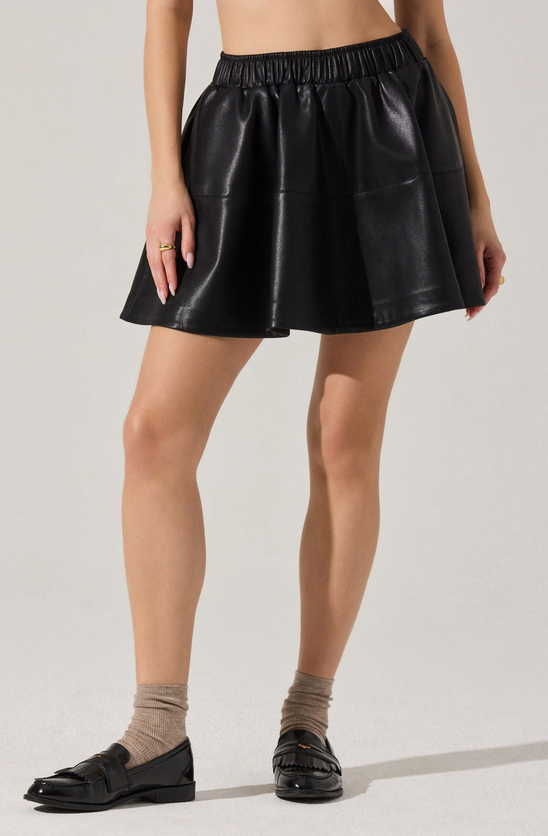 A person is wearing a black faux leather skirt with an elastic waistband, featuring a slight flare and shiny finish. They are also wearing taupe socks and black shiny loafers with fringe details. The person's upper body is not visible, and there are no additional accessories or clothing items shown.