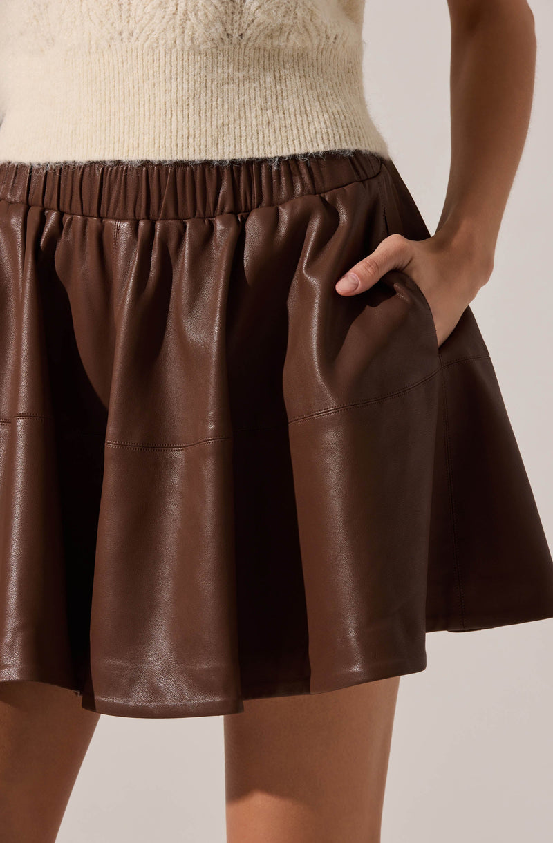 A close-up of a person wearing a beige knitted sweater tucked into a brown, shiny, faux leather skirt with a gathered, elastic waistband and large, voluminous pleats. The person's right hand is resting inside a side pocket of the skirt.