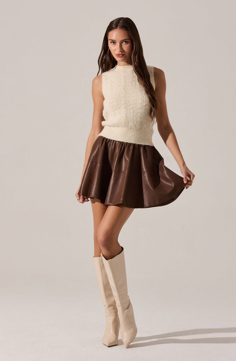 A woman is wearing a sleeveless, knitted cream-colored sweater with a high neckline, a brown satin mini skirt with a gathered waist, and knee-high, pointed-toe cream boots.