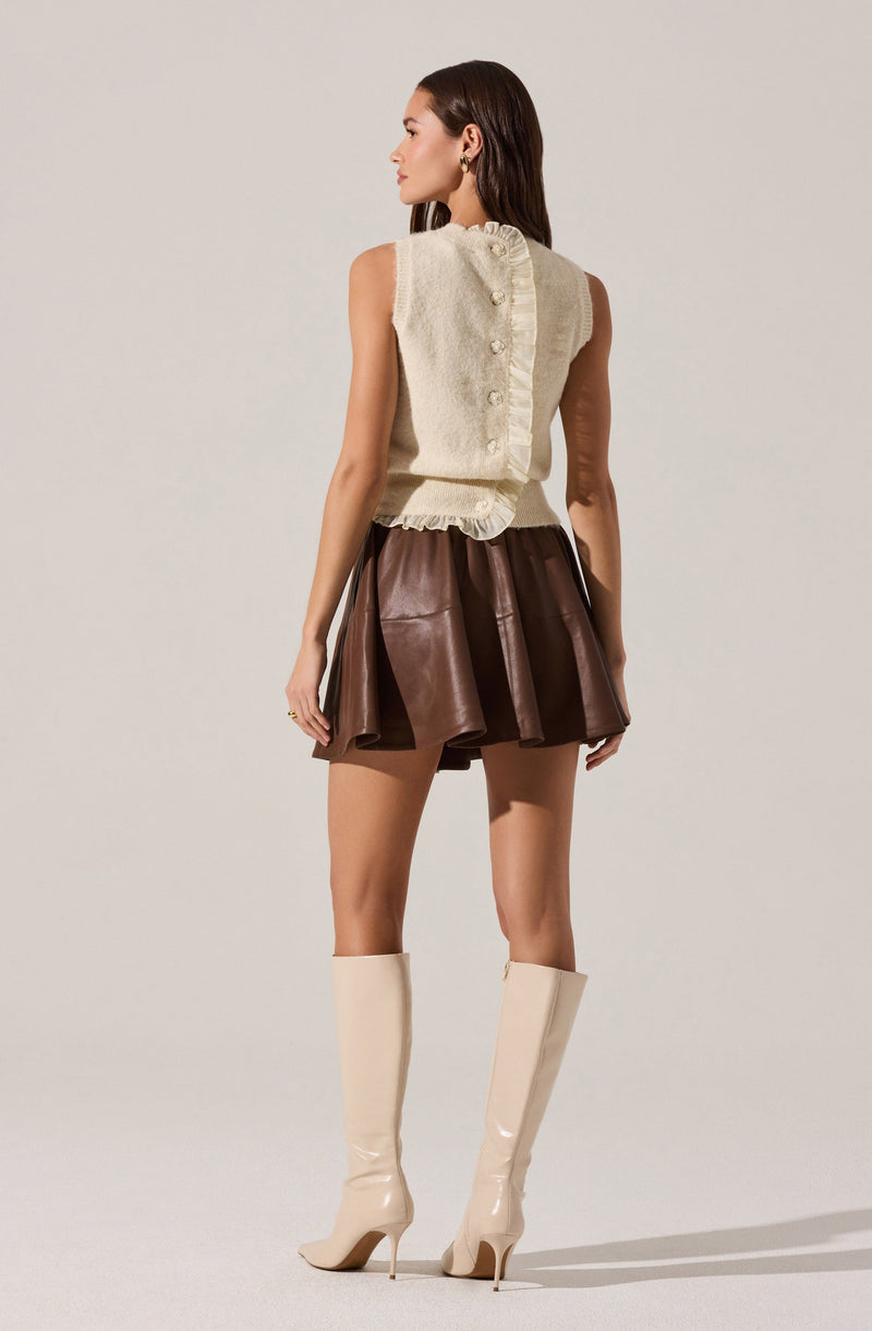 A woman is wearing a sleeveless, cream-colored knitted top with a ruffled trim along the sides and small buttons down the back. She pairs it with a high-waisted, dark brown leather skirt that has a flared, pleated design. She is also wearing knee-high, glossy cream-colored boots with high heels.