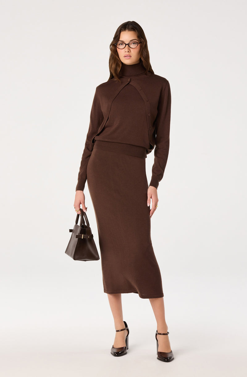 Woman wearing a brown turtleneck sweater and matching midi skirt, holding a brown handbag.